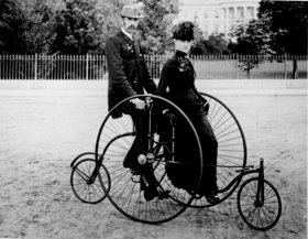 Smartly dressed couple seated on an 1886-model bicycle for two.